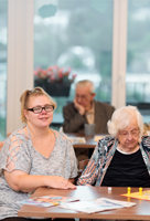 Koordinierende Präsenzkraft sitzt gemeinsam mit einer Seniorin an einem Tisch und spielt Mensch ärgere Dich nicht in ruhiger, freundlicher Atmosphäre.