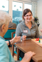 Präsenzkraft sitzt gemeinsam mit einer Seniorin an einem Tisch und spielt Mensch ärgere Dich nicht in ruhiger, freundlicher Atmosphäre.