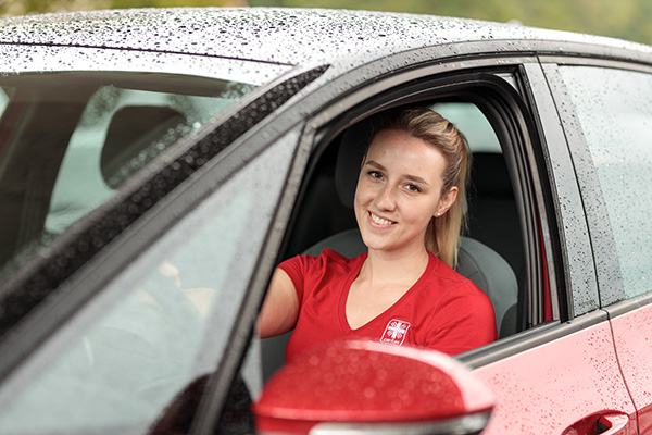 Pflegefachkraft sitzt entspannt im Auto, blickt aus dem Fenster und lächelt in die Kamera – bereit für Pflege mit mehr Zeit und Wertschätzung.
