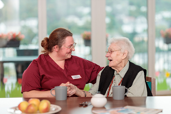 Pflegefachfrau sitzt mit Seniorin am Tisch beim Kaffee – beide lächeln freundlich, positive Pflegesituation.