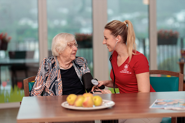 Pflegefachfrau misst einer Seniorin den Blutdruck, beide lächeln sich an – Pflegekräfte stärken und Pflege aktiv gestalten.