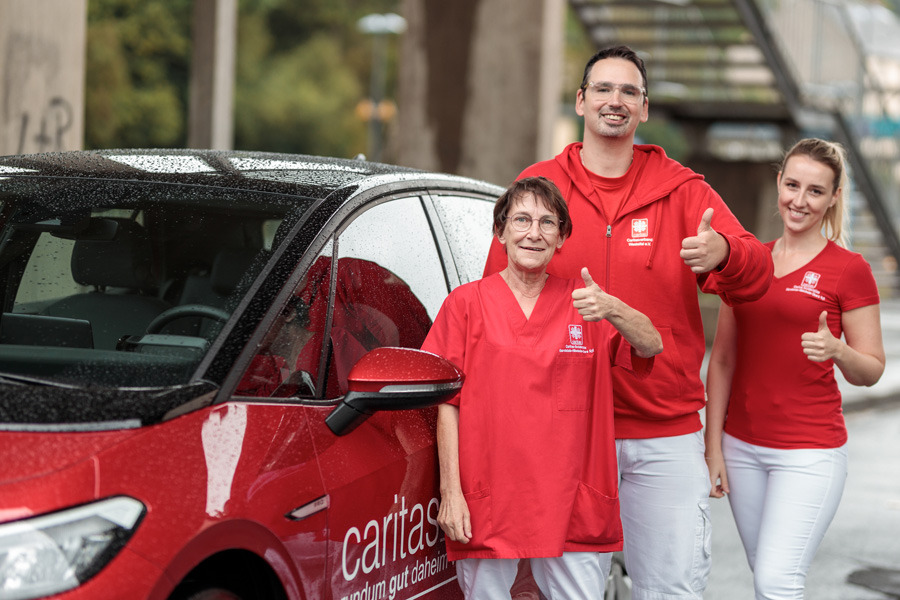 Drei Pflegefachkräfte stehen lachend mit Daumen nach oben am Caritas-Auto – gemeinsam für mehr Flexibilität in der Pflege.