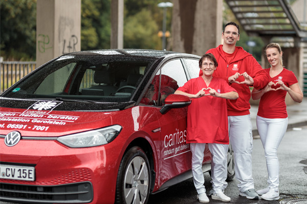 Drei Pflegekräfte am Auto formen jeweils ein Herz mit den Händen – symbolisch für Zusammenhalt im Pflege-Team.