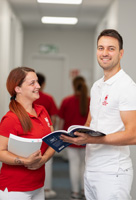Teamleiter lächelt in die Kamera mit Buch, Pflegefachfrau schaut ihn interessiert an.